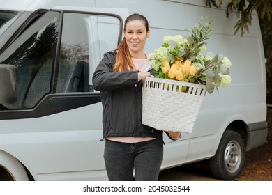 Woman With A Delivery Truck Carries A Large Basket Of Flowers. Deliver Flowers. Supply Garden Plants. Gardener Company Brings Flowers Home. Delivery Woman Carries Flowers