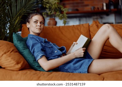 Woman in denim shirt sitting on couch with crossed legs reading a book in cozy home environment - Powered by Shutterstock