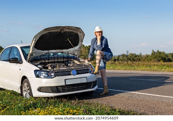 a woman in a denim\
suit, hat and boots stands at the open hood of a broken car on the\
road in a field
