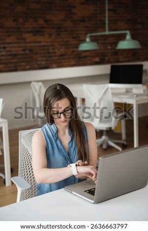 Woman in denim top checking smartwatch while sitting at desk with open laptop in coworking space. Modern, workspace, collaboration, technology, productivity, professional, urban