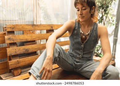 A woman in a denim vest and jeans sits on a wooden bench. She is wearing a necklace and a bracelet - Powered by Shutterstock