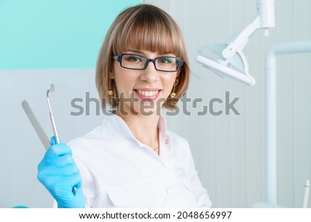 Woman dentist holding tools on background of the dental office in a white coat and glasses smiles, thinks about an idea. Oral health concept