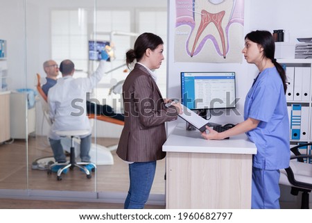 Woman in dentistiry clinic asking information to fill form for theeth treatment from receptionist. Doctor treating senior man in stomatology office. Examination radiograhy.