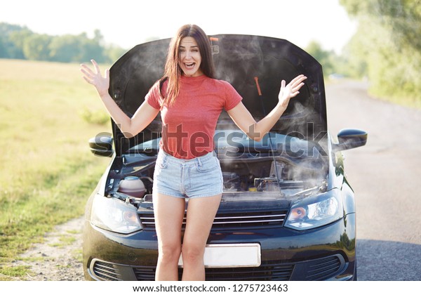 Woman in despair raises hands, stand near brocken\
automobile, smoke from hood, faces technical problem, wears casual\
t shirt and jean shorts, has slender legs, stops on road. Transport\
concept