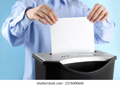Woman destroying sheet of paper with shredder on light blue background, closeup