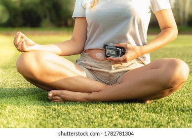 
Woman With Diabetes Is Doing Yoga. The Girl Holds An Insulin Pump In Her Hands. A Woman Is Sitting On The Grass In The Park. Healthy Lifestyle With Diabetes