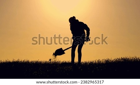 Woman digging pit with shovel dark silhouette in farm field at sunset. Woman digs ground in field against sky lit by sunset. Woman works digging soil with shovel in country field in spring evening