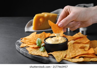 Woman dipping crispy nacho into delicious cheese sauce at black table, closeup. Space for text