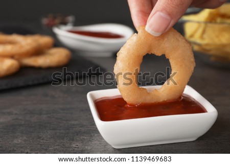 Woman dipping onion ring into hot chili sauce in bowl on table