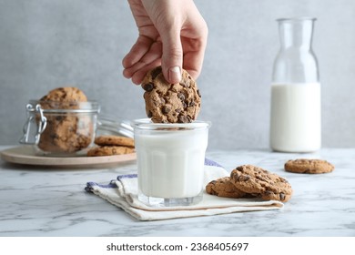 Woman dipping tasty chocolate chip cookie into glass of milk at white marble table, closeup