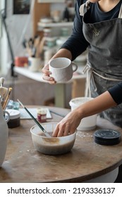 Woman In Dirty Apron Holds White Ceramic Mug Standing At Round Wooden Table On Blurred Background In Workshop. Female Craftsperson Puts Hand Into Bowl With Paint To Work With Handmade Pottery