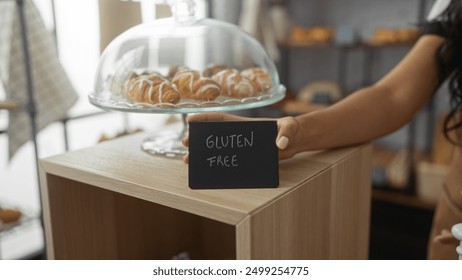 Woman displaying a gluten-free sign in a bakery with pastries under a glass dome, emphasizing the gluten-free options available.