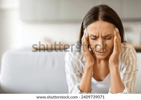 Woman in distress with severe headache, pressing her temples and frowning, young female showing grimace of pain while sitting on couch in bright living room, having signs of stress or migraine