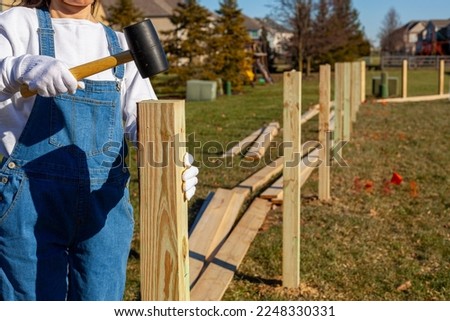 A woman diy enthusiast wearing overalls and safety gloves is installing four by four posts in the perimeter of her backyard. Concept image for fence installation in suburban neighborhood.