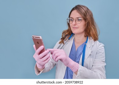 Woman Doctor With An Apple IPhone Phone In A White Uniform On A Blue Studio Background - Moscow, Russia, August 12, 2021