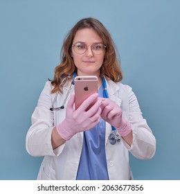 Woman Doctor With An Apple IPhone Phone In A White Uniform On A Blue Studio Background - Moscow, Russia, August 12, 2021