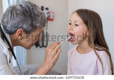 Woman doctor examining little girl mouth at office. Senior doctor at hospital checking the sore throat of a young patient. Pediatrician checking tonsils of a little patient in hospital room.
