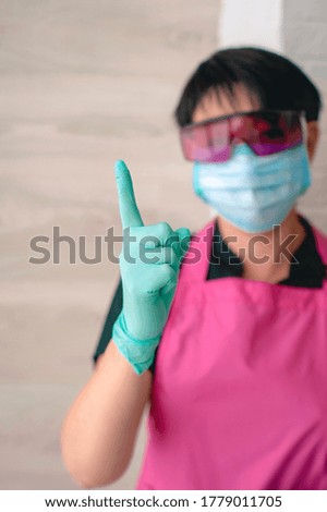 woman doctor in glasses, apron, rubber gloves and mask showing sign - finger up, aware. Aware, sign, symbol 