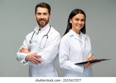 Woman doctor holding clipboard and posing with her male colleague with stethoscope with happy faces while standing isolated on grey background 