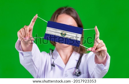 Woman doctor in a medical coat holds a medical mask with of the El Salvador flag on a green isolated background
