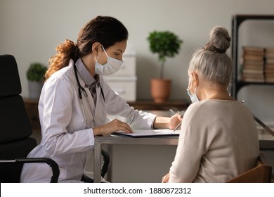 Woman Doctor In Medical Facemask Talk Consult Elderly Patient About Health Insurance Deal With Hospital. Female GP In Facial Mask Prescribe Treatment, Consider Results With Senior Client In Clinic.