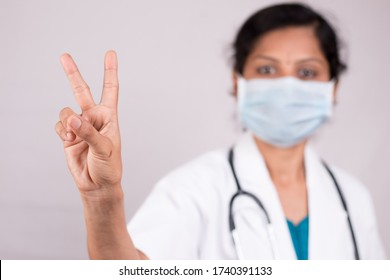 Woman Doctor With Medical Mask In Uniform Showing V Gesture On Isolated Background - Concept Of Victory Salute Or Peace Gesture By Doctor During Covid-19 Or Coronavirus Pandemic.