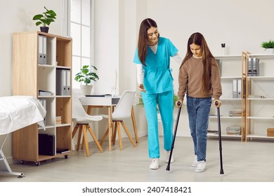 Woman doctor or nurse in a medical scrubs uniform helps a little kid child patient girl walk with crutches at the clinic during a rehabilitation course after her leg injury - Powered by Shutterstock