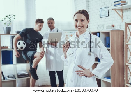 Woman doctor stands in the office and posing in a white coat. Male soccer player with knee injury stands near doctor with soccer ball. 