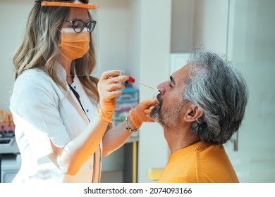 Woman Doctor Wearing Lab Coat, Protective Face Mask, Face Shield and Orange Gloves Using Cotton Swab to Test her Mature Male Patient on Possible Coronavirus Infection at Medical Clinic