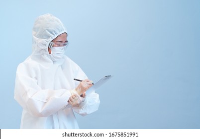 A Woman Doctor Wearing Personal Protective Equipment  Or Ppe Including White Suit, Mask And Gloves Is Writing The Documents On The Blue Background. Coronavirus, Covid 19, Medical, Healthcare Concept