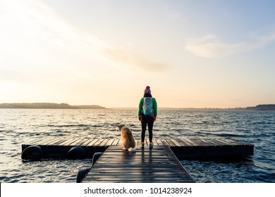 Frauen mit Hund genießen Sonnenaufgang und See, entspannen auf der Brücke. Wanderer oder Touristen, die einen schönen Blick auf den Morgen mit Hund Freund, inspirierende Landschaft am Strand. Friedliche Menschen und ruhiges Konzept.