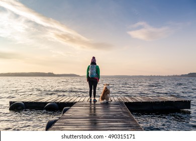 Woman with dog enjoy sunrise and lake, relaxing on bridge. Hiker or tourist looking at beautiful morning view with dog friend, inspirational landscape on beach. Peaceful people and serene concept.