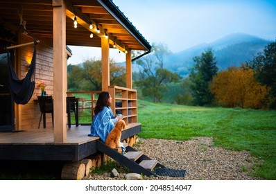  Woman with dog resting on the terrace mountain chalet at sunset.
