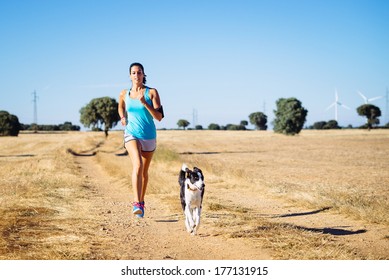 Woman and dog running in country side dirt track. Female runner exercising and training with her pet for cross race. Fitness girl on summer rural landscape.