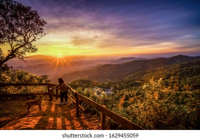 Woman and dog standing on the top of the hill in sunset over village and beautiful mountains background - Shutterstock ID 1629455059