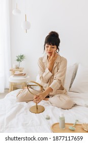 Woman Doing Beauty Self Care Sitting At Home. Woman Sitting On Bed Applying Cosmetics Looking At Small Mirror.