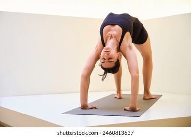 A woman is doing chakrasana or wheel yoga pose on a mat. The pose is a backbend. Concept of focus and determination, as the woman is fully engaged in her yoga practice. - Powered by Shutterstock