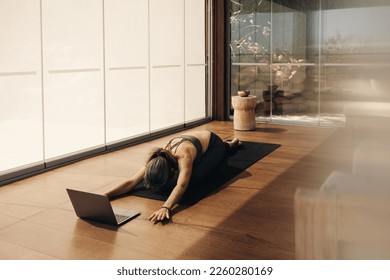 Woman doing a child's pose exercise during an online yoga class at home. Senior woman practicing a kneeling asana on a yoga mat. Mature woman following a virtual workout tutorial on a laptop. - Powered by Shutterstock