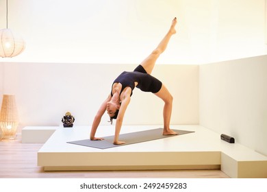 A woman is doing Eka Pada Chakrasana or one legged wheel yoga pose on a mat. The pose is a backbend. Concept of focus and determination, as the woman is fully engaged in her yoga practice. - Powered by Shutterstock