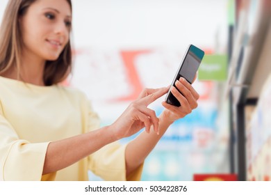 Woman Doing Grocery Shopping At The Supermarket And Using A Smart Phone, She Is Checking Offers And Discounts