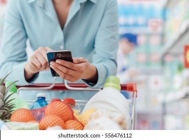 Woman Doing Grocery Shopping At The Supermarket, She Is Leaning On The Shopping Cart And Connecting With Her Phone, Apps And Retail Concept