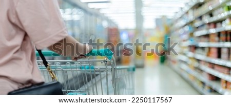Woman doing grocery shopping at the supermarket, pushing a shopping cart. Shopping cart in supermarket aisle, copy space. Closeup of a woman shopper with a modern shopping cart at supermarket