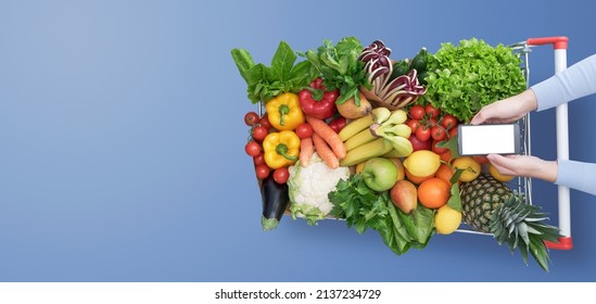 Woman doing grocery shopping and using her smartphone, her shopping cart is full of fresh vegetables and fruits, top view - Shutterstock ID 2137234729