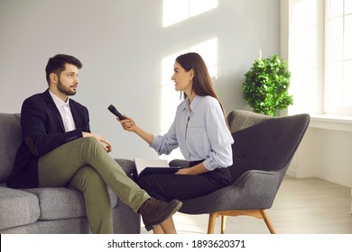 Woman doing interview with male celebrity, opinion leader or business influencer. Female newspaper journalist or TV host sitting in studio, holding microphone and asking famous young man questions