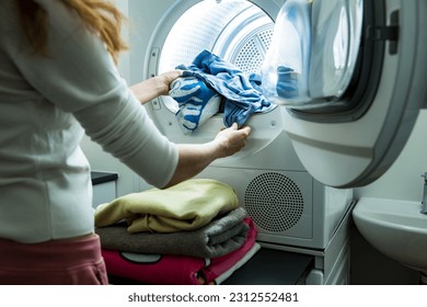 Woman doing laundry. Dryer machine in a Landry room at home drying clothes. Housewife unloading dryer and folding clean and dry linen. Household chores concept 