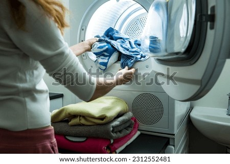 Woman doing laundry. Dryer machine in a Landry room at home drying clothes. Housewife unloading dryer and folding clean and dry linen. Household chores concept 