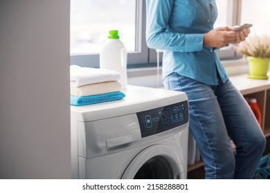 Woman Doing Laundry At Home Using A Smart Washing Machine, She Is Controlling The Smart Washer From Her Smartphone