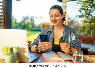 Woman Is Doing Online Purchases. Girl Is Doing Shopping Using Phone And Credit Card Sitting On The Patio In Summer.