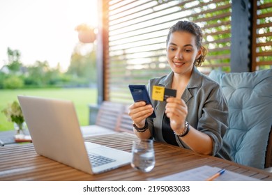 Woman Is Doing Online Purchases. Girl Is Doing Shopping Using Phone And Credit Card Sitting On The Patio In Summer.