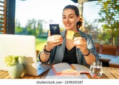 Woman Is Doing Online Purchases. Girl Is Doing Shopping Using Phone And Credit Card Sitting On The Patio In Summer.